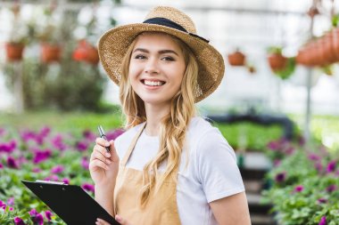 Blonde woman holding clipboard by flowers in greenhouse clipart