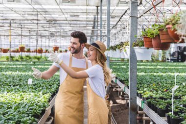 Blonde woman and handsome man in aprons talking among flowers in glasshouse clipart