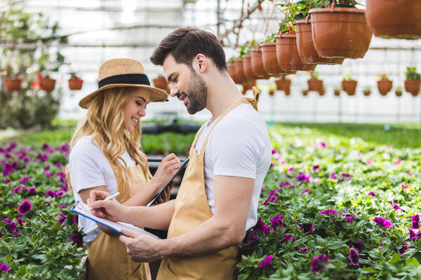 Couple of gardeners holding clipboards by flowers in greenhouse