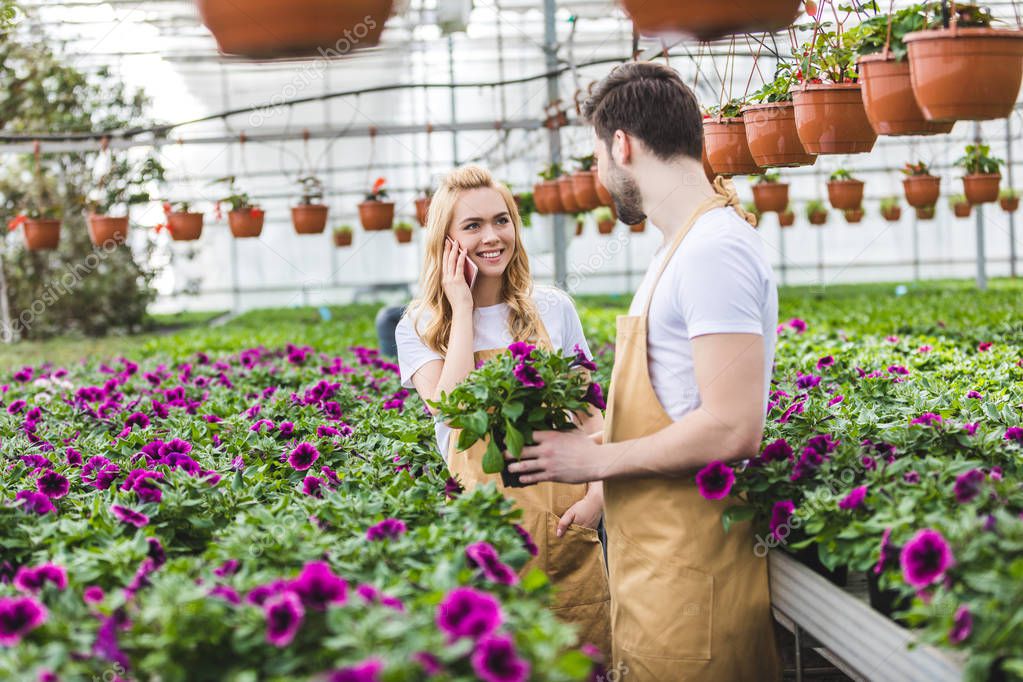 Young male gardener holding pot with flowers while woman talking on phone