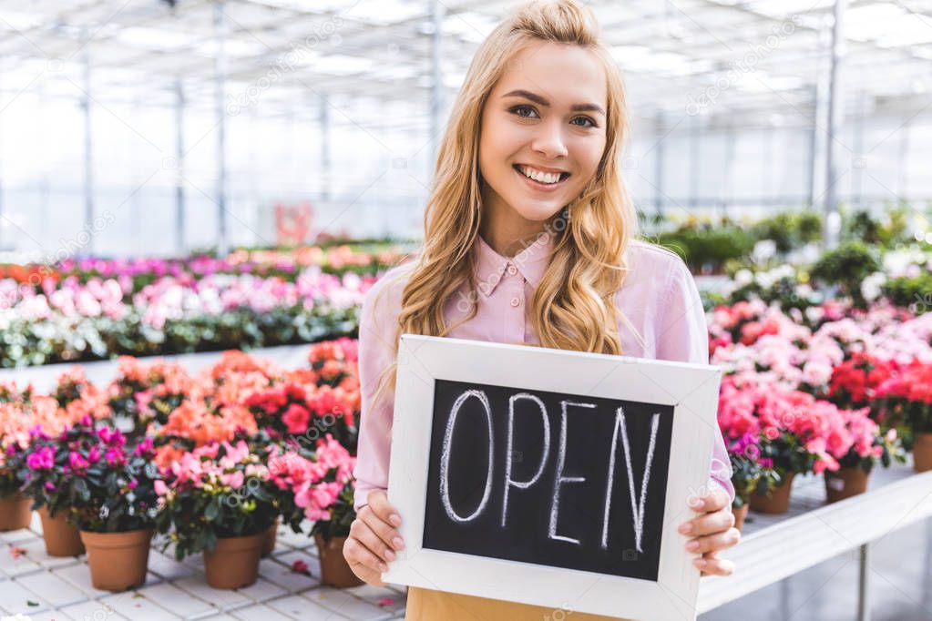 Attractive female gardener holding Open board by flowers in glasshouse