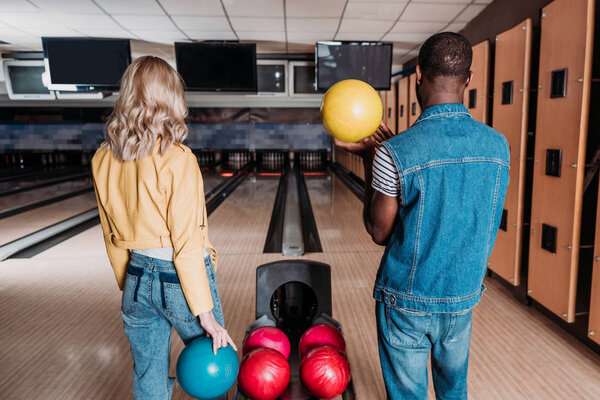 multiethnic couple with bowling balls standing in front of alleys at club