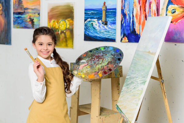 smiling kid holding painting brush and canvas in workshop of art school