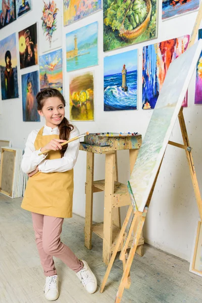 Uśmiechający Się Dziecko Stojąc Pędzlem Pobliżu Canvas Warsztacie Szkoły Sztuki — Zdjęcie stockowe