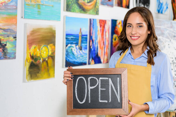 happy female artist holding signboard with word open in workshop
