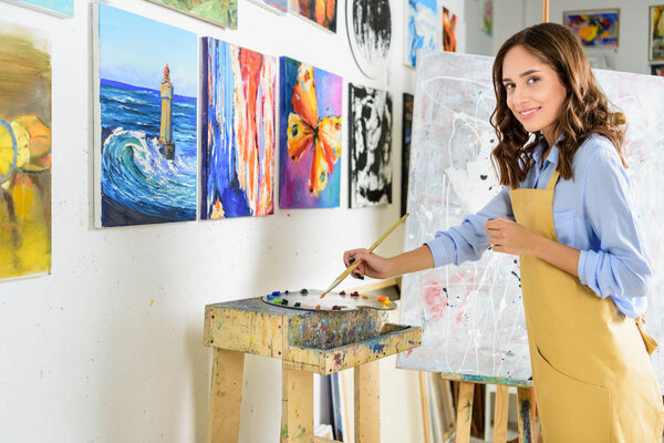 вид сбоку на красивую художницу, которая берет краску из палитры в мастерской
