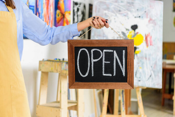 cropped image of female artist holding signboard with word open in workshop