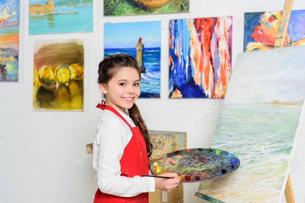 preteen kid holding palette near canvas in workshop of art school
