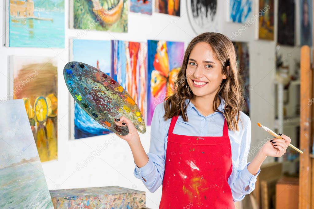 smiling beautiful female artist holding painting brush and palette in workshop