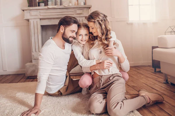 Glückliche junge Familie mit einem Kind, das zu Hause auf dem Teppich sitzt und sich umarmt — Stockfoto