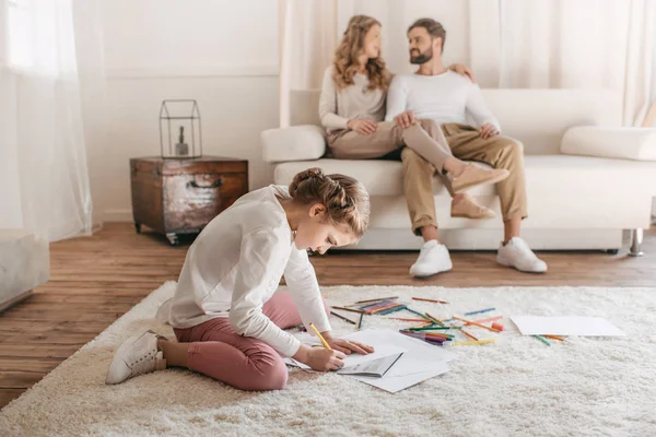 Little girl drawing and sitting on floor, parents sitting on sofa behind — Stock Photo