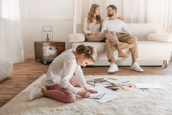 Petite fille dessin et assis sur le sol, parents assis sur le canapé derrière — Photo de stock