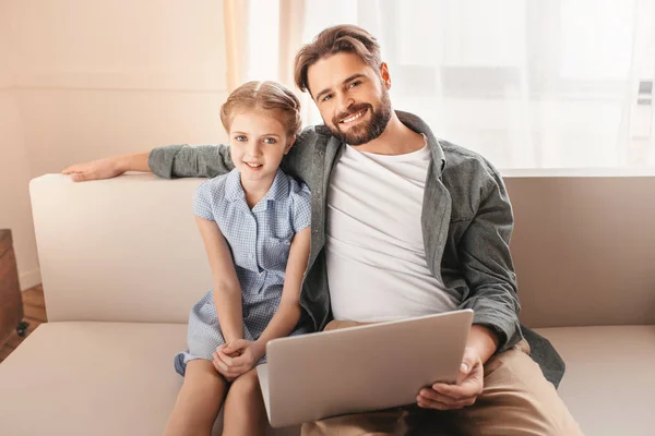 Feliz padre e hija sentados en el sofá y utilizando el ordenador portátil en casa - foto de stock