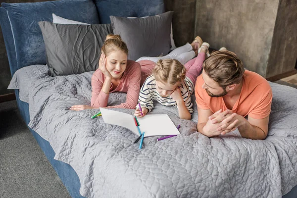 Happy parents lying on bed and looking at smiling daughter drawing — Stock Photo