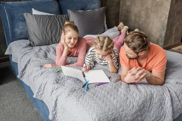 Padres felices acostados en la cama y mirando al dibujo sonriente de la hija - foto de stock