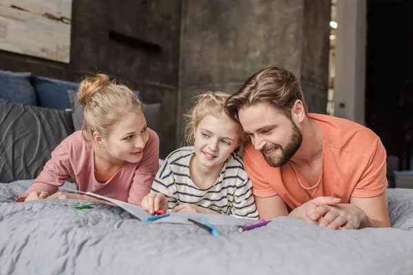 Parents heureux couchés sur le lit et regardant la fille souriante dessin — Photo de stock