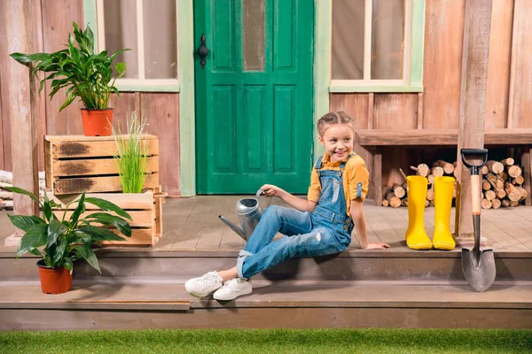 Adorable niña sonriente sentada en el porche con regadera y mirando hacia otro lado - foto de stock