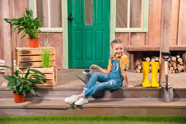 Adorable smiling little girl sitting on porch with watering can and looking away — Stock Photo