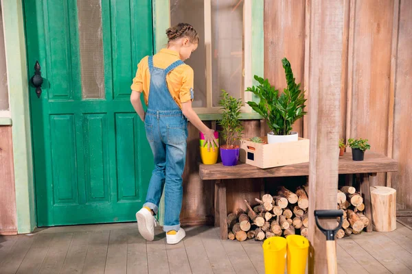 Back view of little girl in denim overalls standing on porch with flower pots — Stock Photo