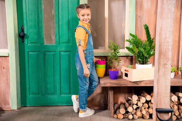 Adorable niña de pie en el porche con macetas de colores y sonriendo a la cámara - foto de stock