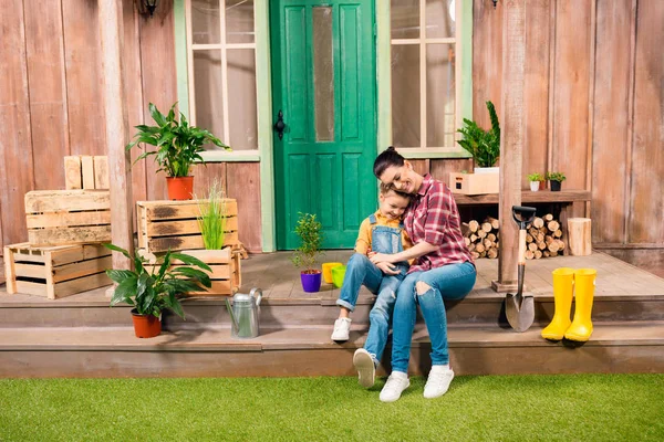 Feliz madre e hija sentadas y cogidas de la mano en el porche con plantas en maceta - foto de stock