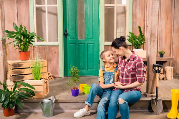 Feliz madre e hija sentadas y cogidas de la mano en el porche con plantas en maceta - foto de stock