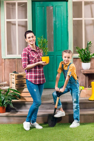 Mãe sorridente com planta em vaso e filha com pá de jardim em pé no alpendre — Fotografia de Stock
