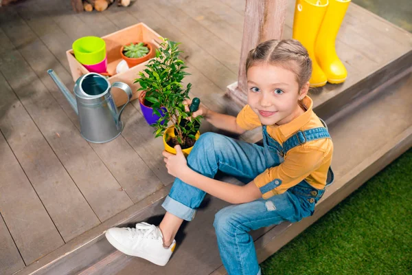 High angle view of adorable little girl sitting on porch and cultivating green plant in pot — Stock Photo