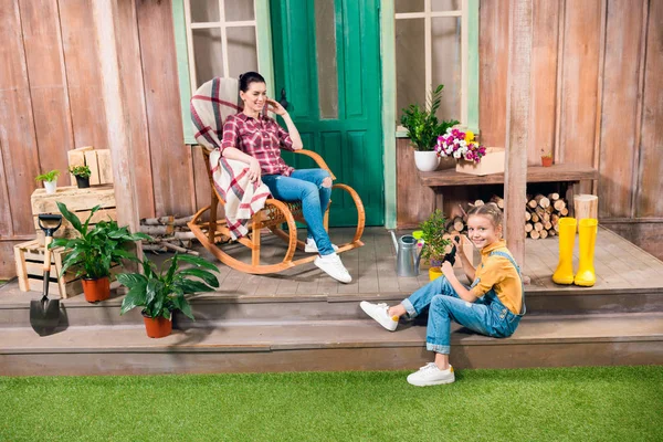 Mãe feliz sentada na cadeira de balanço e sorrindo filha cultivando planta no alpendre — Fotografia de Stock