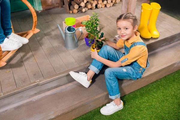 Vue grand angle de adorable petite fille assise sur le porche et cultivant la plante verte en pot — Photo de stock