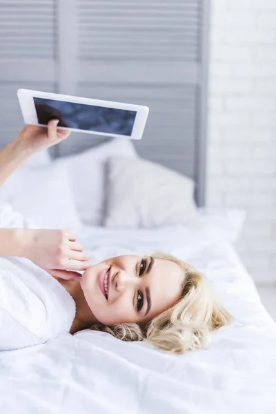 Привлекательная блондинка держит цифровой планшет с чистым экраном и улыбается на камеру в спальне — стоковое фото