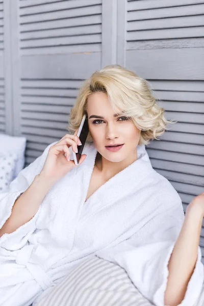 Beautiful blonde girl looking at camera while talking by smartphone on bed — Stock Photo
