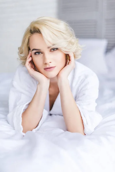 Beautiful blonde woman in bathrobe looking at camera while lying on bed — Stock Photo