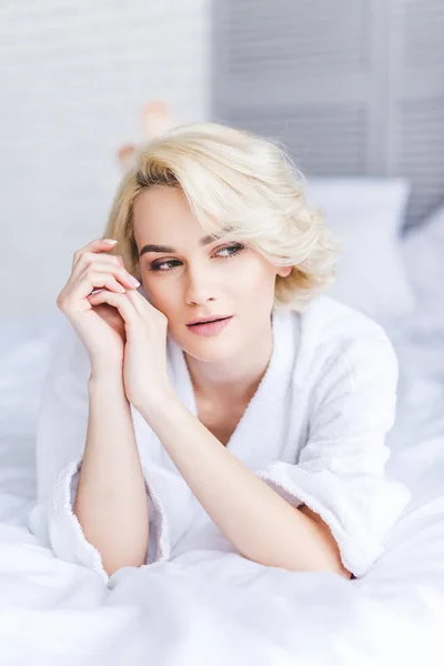 Задумчивая привлекательная блондинка в халате смотрит в сторону в спальне — стоковое фото