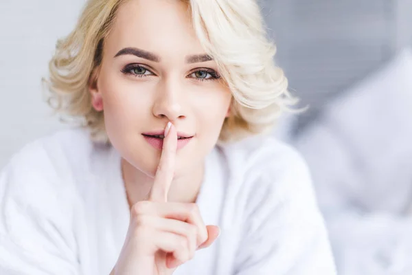 Beautiful blonde woman in bathrobe gesturing for silence and looking at camera — Stock Photo