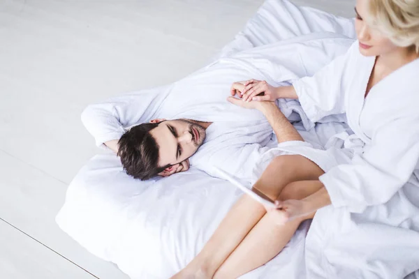 Молодая пара в халатах, держась за руки, пока девушка использует цифровой планшет на кровати — стоковое фото