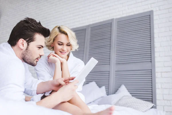 Sourire jeune couple en peignoirs à l'aide d'une tablette numérique sur le lit — Photo de stock