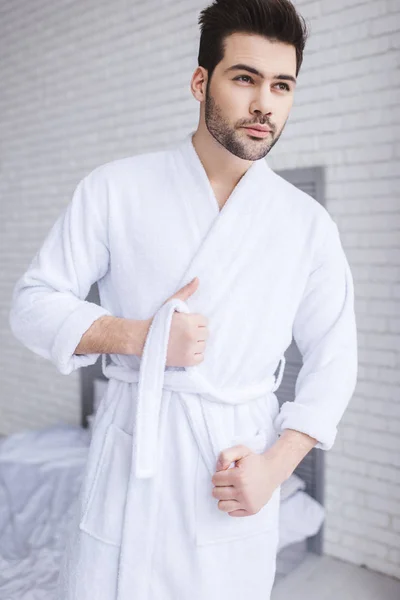 Handsome young man tying belt of bathrobe and looking away at home — Stock Photo