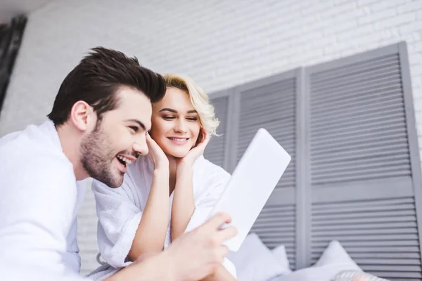 Feliz pareja joven en albornoces usando tableta digital en el dormitorio - foto de stock