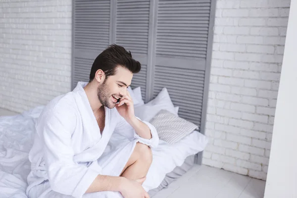 Улыбающийся молодой человек в халате разговаривает по смартфону, сидя на кровати — стоковое фото