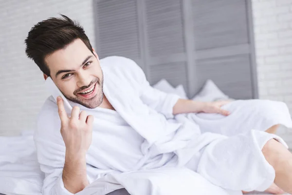 Handsome young man in bathrobe talking by smartphone and smiling at camera — Stock Photo