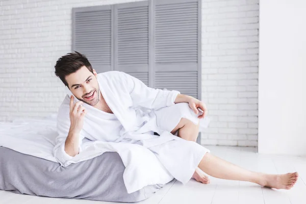 Joven guapo en albornoz hablando por teléfono inteligente y mirando a la cámara - foto de stock