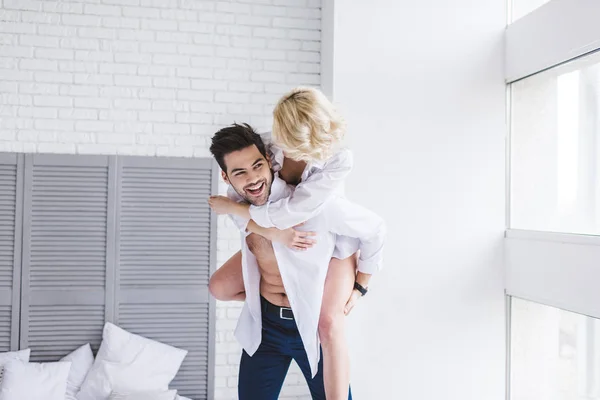 Hermosa feliz joven pareja en blanco camisas piggybacking en dormitorio - foto de stock