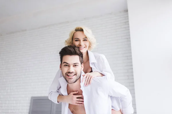 Hermosa feliz joven pareja piggybacking y sonriendo a la cámara en casa - foto de stock