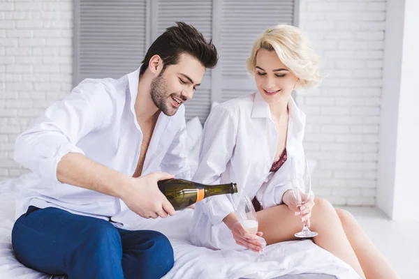 Красивый улыбающийся молодой человек наливает шампанское в бокалы, сидя с девушкой на кровати — стоковое фото