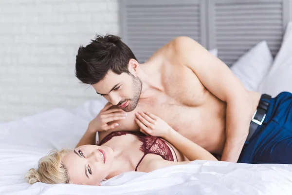 Seductive young woman in underwear smiling at camera while lying with boyfriend on bed — Stock Photo