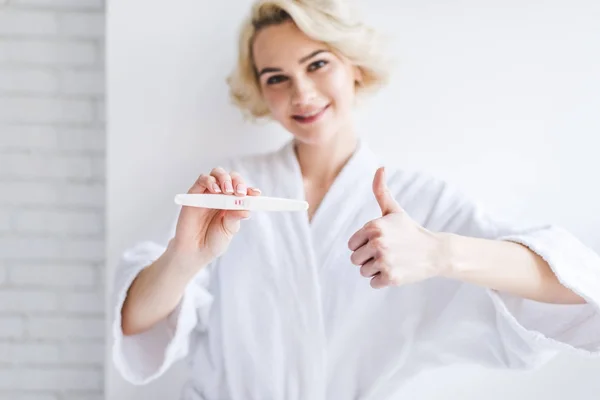 Sorridente giovane donna con test di gravidanza positivo e mostrando pollice in su — Foto stock