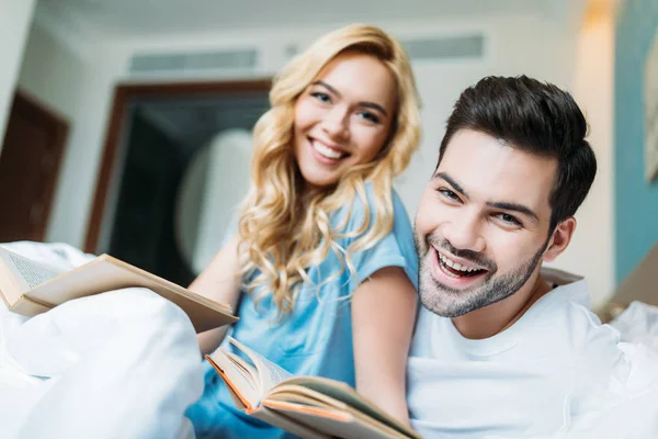 Lächelndes Paar mit Büchern im Bett, die in die Kamera schauen — Stockfoto