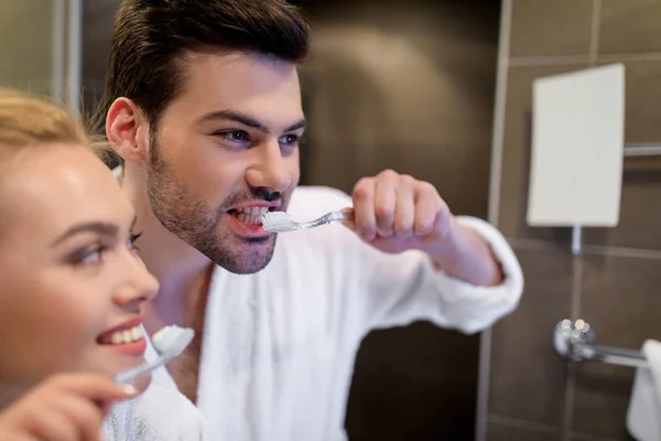 Sonriente pareja cepillándose los dientes y mirando al espejo - foto de stock