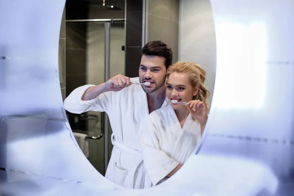 Pareja sonriente en albornoces cepillándose los dientes y mirando al espejo - foto de stock